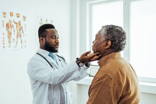 An ENT specialist examines an older male patient’s throat in a medical office, gently holding his chin while assessing his neck.